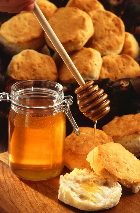 A glass jar of honey with a wooden dipper drizzling honey onto a split biscuit